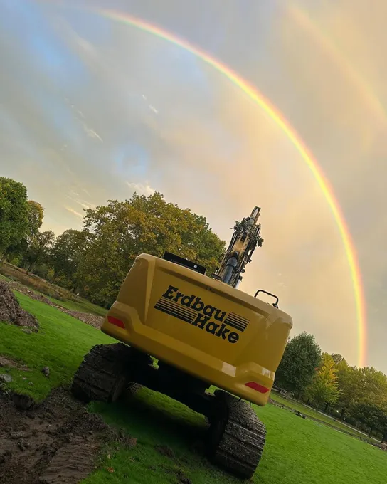 Gelber Bagger mit Erdbau-Hake-Logo unter einem doppelten Regenbogen